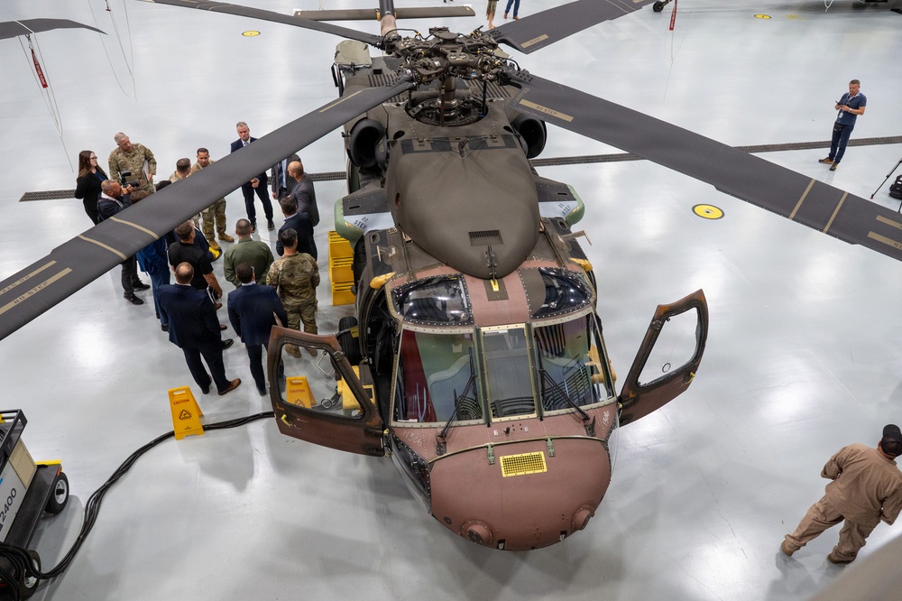 An overhead view of a military helicopter, likely a Sikorsky UH-60 Black Hawk variant, displayed inside a large hangar facility. A group of mixed civilian and military personnel are gathered around the aircraft, suggesting an official demonstration, briefing, or inspection event. The helicopter appears to be in a partially disassembled or maintenance state, with exposed components visible.