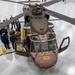 An overhead view of a military helicopter, likely a Sikorsky UH-60 Black Hawk variant, displayed inside a large hangar facility. A group of mixed civilian and military personnel are gathered around the aircraft, suggesting an official demonstration, briefing, or inspection event. The helicopter appears to be in a partially disassembled or maintenance state, with exposed components visible.