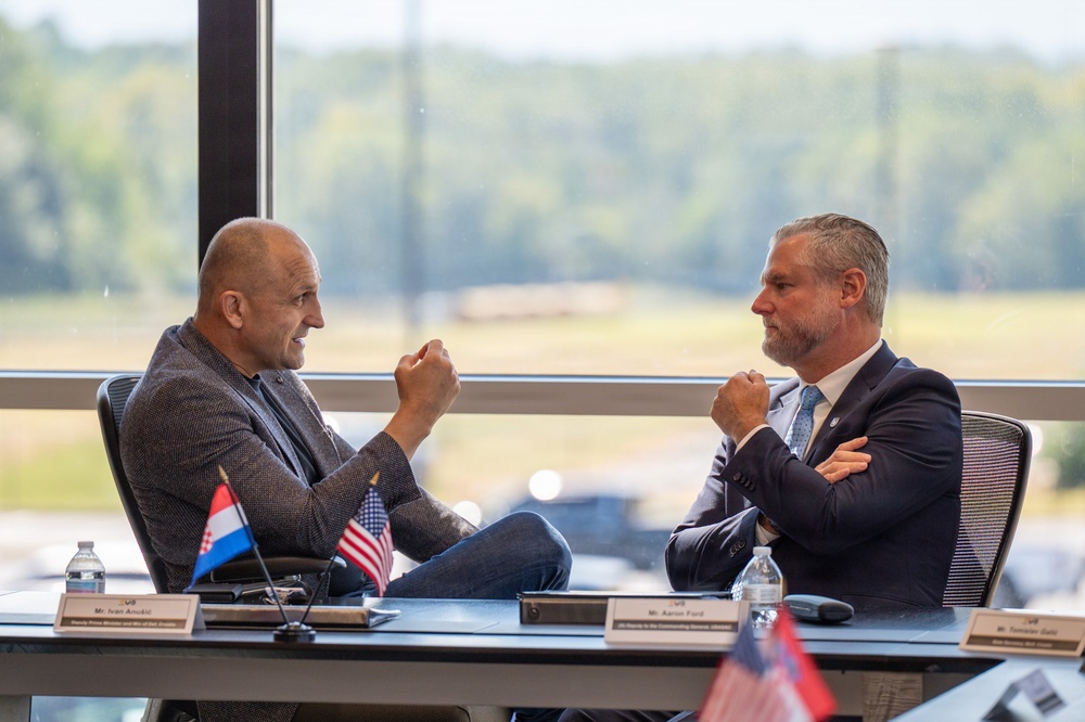 Two men are engaged in a formal bilateral meeting at a conference table, with name placards and national flags visible. The man on the left is identified as Mr. Ivan Anušić, Deputy Prime Minister and Minister of Defence of Croatia, and the man on the right is identified as Mr. Aaron Ford, with a third placard partially visible. The setting appears to be a modern conference room with large windows overlooking a green landscape.