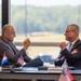Two men are engaged in a formal bilateral meeting at a conference table, with name placards and national flags visible. The man on the left is identified as Mr. Ivan Anušić, Deputy Prime Minister and Minister of Defence of Croatia, and the man on the right is identified as Mr. Aaron Ford, with a third placard partially visible. The setting appears to be a modern conference room with large windows overlooking a green landscape.