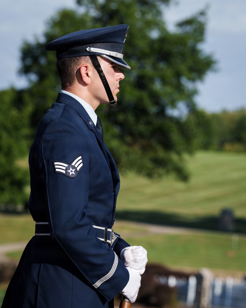 A United States Air Force Senior Airman stands at attention in dress blues uniform, appearing to participate in a formal military ceremony. The service member is photographed in profile view, wearing white gloves and holding what appears to be a rifle or ceremonial staff. The setting appears to be a military cemetery or memorial grounds, visible in the blurred background.