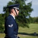 A United States Air Force Senior Airman stands at attention in dress blues uniform, appearing to participate in a formal military ceremony. The service member is photographed in profile view, wearing white gloves and holding what appears to be a rifle or ceremonial staff. The setting appears to be a military cemetery or memorial grounds, visible in the blurred background.