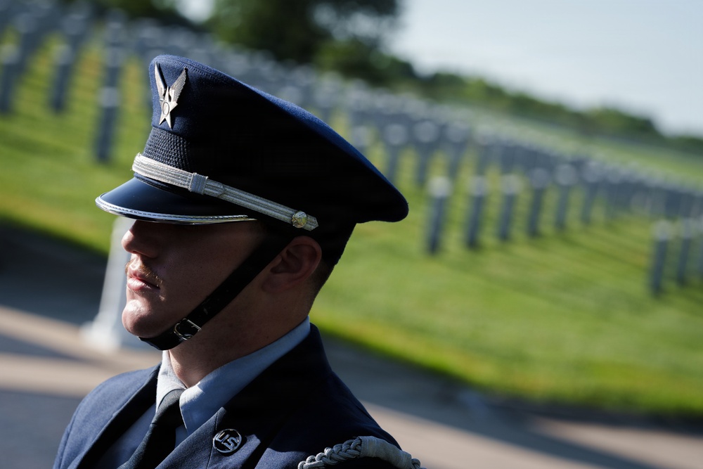 A U.S. Air Force service member in dress uniform stands at attention in what appears to be a military cemetery. The subject is photographed in profile with rows of white grave markers visible and blurred in the background. The image conveys a solemn ceremonial context, likely a memorial or funeral honors detail.