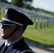 A U.S. Air Force service member in dress uniform stands at attention in what appears to be a military cemetery. The subject is photographed in profile with rows of white grave markers visible and blurred in the background. The image conveys a solemn ceremonial context, likely a memorial or funeral honors detail.