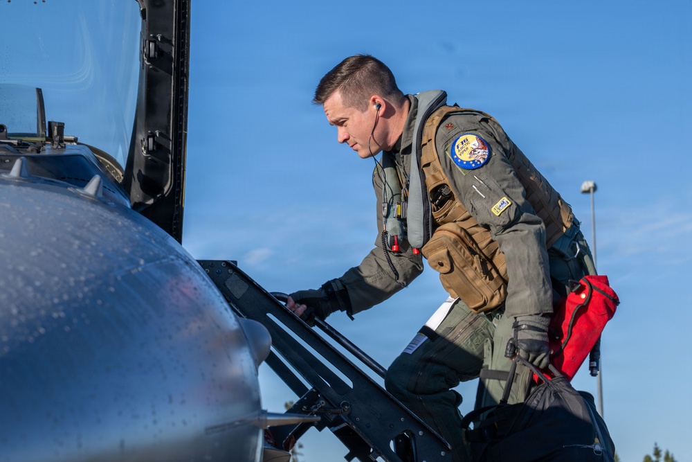 A military pilot in full flight gear is climbing into the cockpit of a jet aircraft, likely an F-16 based on the visible patch on his vest. He is wearing an olive-green flight suit, a tactical survival vest, and black gloves, and is carrying a red bag and a dark flight bag. The image is taken outdoors under a clear blue sky.
