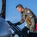 A military pilot in full flight gear is climbing into the cockpit of a jet aircraft, likely an F-16 based on the visible patch on his vest. He is wearing an olive-green flight suit, a tactical survival vest, and black gloves, and is carrying a red bag and a dark flight bag. The image is taken outdoors under a clear blue sky.