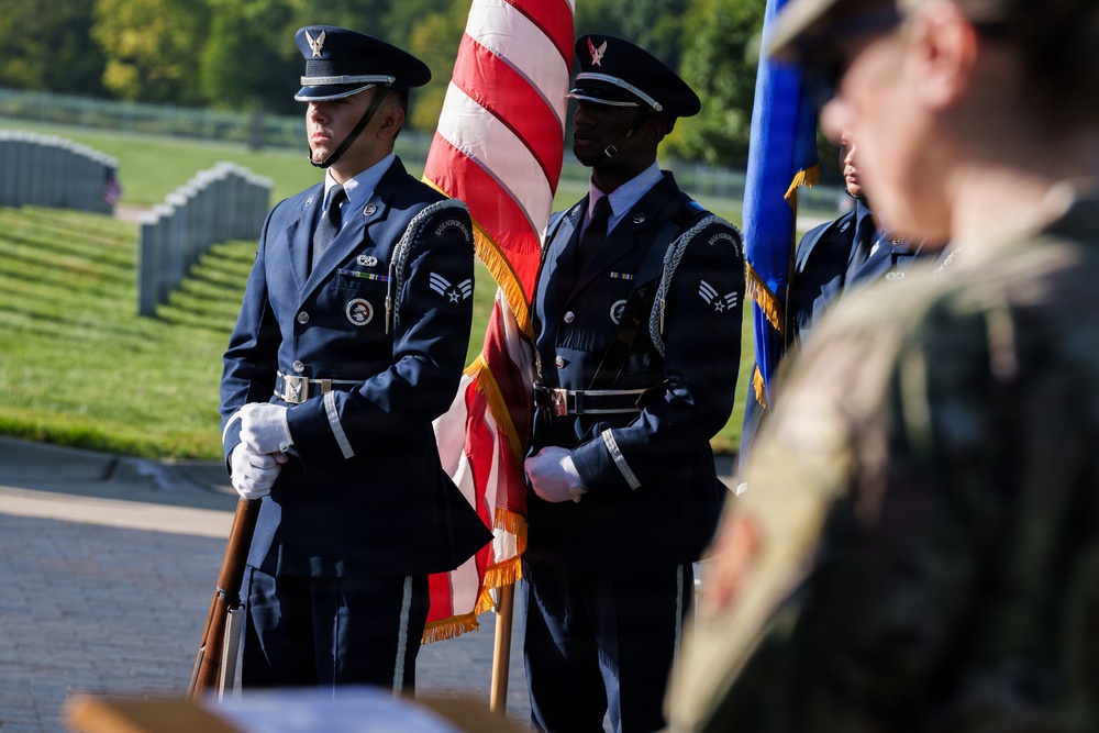 A U.S. Air Force Honor Guard color guard unit stands at attention during a formal military ceremony, likely at a national cemetery based on the grave markers visible in the background. The airmen are dressed in ceremonial dress blues and are carrying the American flag and what appears to be an Air Force flag. The setting and attire suggest this is a memorial or commemorative event.