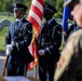 A U.S. Air Force Honor Guard color guard unit stands at attention during a formal military ceremony, likely at a national cemetery based on the grave markers visible in the background. The airmen are dressed in ceremonial dress blues and are carrying the American flag and what appears to be an Air Force flag. The setting and attire suggest this is a memorial or commemorative event.