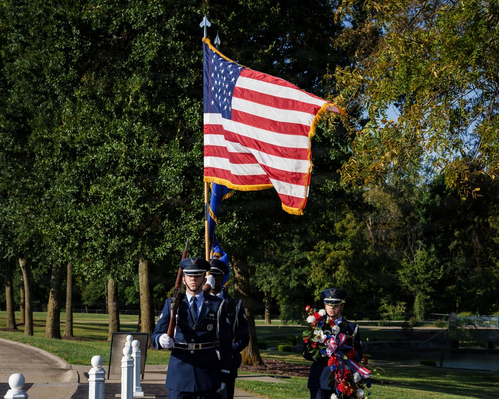A military honor guard ceremony is taking place outdoors in a park-like setting. Personnel in U.S. Air Force dress blue uniforms are carrying the American flag and a service flag, while another member holds a floral wreath. The scene appears to be a formal commemorative or memorial ceremony.