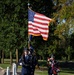 A military honor guard ceremony is taking place outdoors in a park-like setting. Personnel in U.S. Air Force dress blue uniforms are carrying the American flag and a service flag, while another member holds a floral wreath. The scene appears to be a formal commemorative or memorial ceremony.