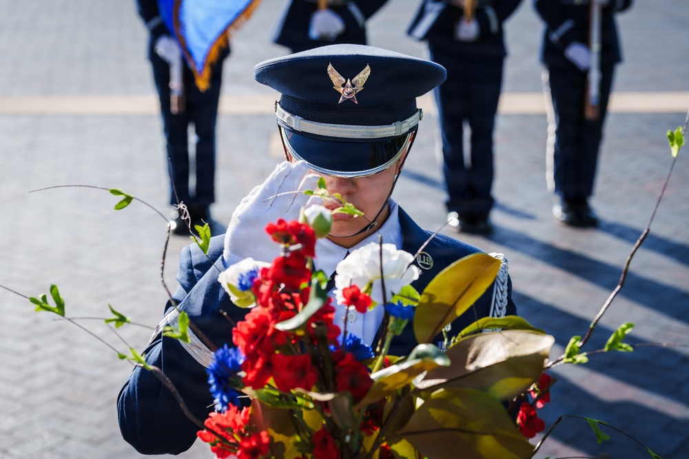 A uniformed military service member, appearing to be from the U.S. Air Force based on the distinctive blue dress uniform and cap insignia, is saluting or bowing their head near a floral arrangement of red, white, and blue flowers. The ceremony appears to be a formal wreath or floral laying event, likely a memorial or commemorative occasion. In the background, additional uniformed personnel are visible, along with what appears to be a color guard carrying flags.