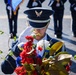 A uniformed military service member, appearing to be from the U.S. Air Force based on the distinctive blue dress uniform and cap insignia, is saluting or bowing their head near a floral arrangement of red, white, and blue flowers. The ceremony appears to be a formal wreath or floral laying event, likely a memorial or commemorative occasion. In the background, additional uniformed personnel are visible, along with what appears to be a color guard carrying flags.