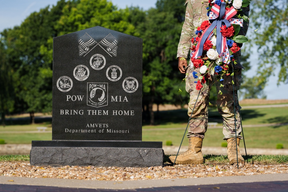 A military service member in camouflage uniform stands beside a black granite POW/MIA memorial monument holding a red, white, and blue floral wreath. The monument reads 'POW MIA - BRING THEM HOME' and is dedicated by AMVETS Department of Missouri. The setting appears to be an outdoor memorial or cemetery grounds during a commemorative ceremony.