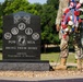 A military service member in camouflage uniform stands beside a black granite POW/MIA memorial monument holding a red, white, and blue floral wreath. The monument reads 'POW MIA - BRING THEM HOME' and is dedicated by AMVETS Department of Missouri. The setting appears to be an outdoor memorial or cemetery grounds during a commemorative ceremony.