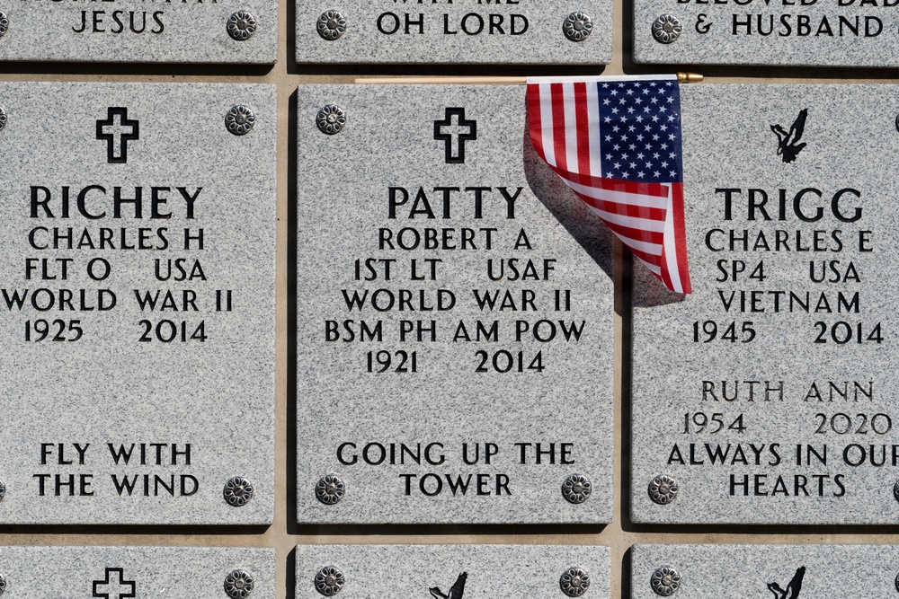The image shows three military memorial grave markers at what appears to be a national cemetery. The center marker has a small American flag placed on it. All three markers honor veterans from different conflicts, including World War II and Vietnam.