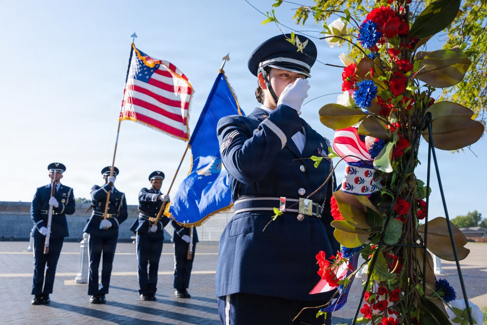 A U.S. Air Force honor guard ceremony is taking place outdoors, likely at a memorial or commemorative event. A female airman in dress blues is rendering a salute near a floral wreath arrangement featuring red, white, and blue flowers. Additional honor guard members stand in formation behind her, carrying the American flag and what appears to be an Air Force flag.