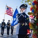 A U.S. Air Force honor guard ceremony is taking place outdoors, likely at a memorial or commemorative event. A female airman in dress blues is rendering a salute near a floral wreath arrangement featuring red, white, and blue flowers. Additional honor guard members stand in formation behind her, carrying the American flag and what appears to be an Air Force flag.