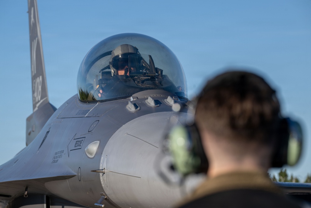 A military fighter jet, identifiable as an F-16 Fighting Falcon, is shown on a flight line with a pilot seated in the cockpit wearing a flight helmet. A ground crew member wearing hearing protection (ear defenders) is visible in the foreground, facing the aircraft. The scene appears to depict pre-flight or post-flight ground operations.