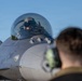 A military fighter jet, identifiable as an F-16 Fighting Falcon, is shown on a flight line with a pilot seated in the cockpit wearing a flight helmet. A ground crew member wearing hearing protection (ear defenders) is visible in the foreground, facing the aircraft. The scene appears to depict pre-flight or post-flight ground operations.