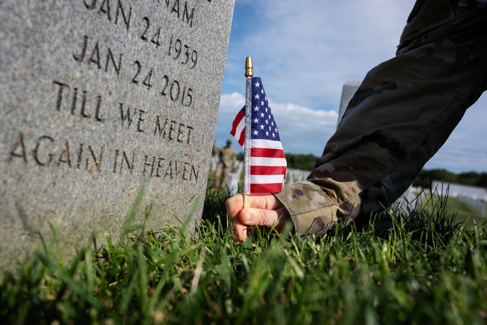 A person in military camouflage uniform is placing a small American flag into the grass at the base of a military gravestone in a cemetery. The headstone bears the dates JAN 24 1939 and JAN 24 2015, along with the inscription 'TILL WE MEET AGAIN IN HEAVEN.' The scene appears to be a Memorial Day or similar commemorative flag-placing ceremony at a national cemetery.