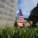 A person in military camouflage uniform is placing a small American flag into the grass at the base of a military gravestone in a cemetery. The headstone bears the dates JAN 24 1939 and JAN 24 2015, along with the inscription 'TILL WE MEET AGAIN IN HEAVEN.' The scene appears to be a Memorial Day or similar commemorative flag-placing ceremony at a national cemetery.