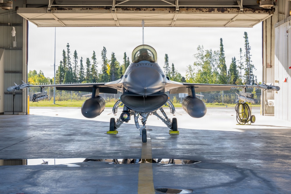 A front-facing view of an F-16 Fighting Falcon jet fighter parked inside a military hangar with the large bay doors open. The aircraft is equipped with wing-mounted munitions and external fuel tanks, positioned on chocks with landing gear deployed. The background reveals a tree line and open airfield consistent with a northern or boreal climate base.