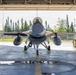 A front-facing view of an F-16 Fighting Falcon jet fighter parked inside a military hangar with the large bay doors open. The aircraft is equipped with wing-mounted munitions and external fuel tanks, positioned on chocks with landing gear deployed. The background reveals a tree line and open airfield consistent with a northern or boreal climate base.