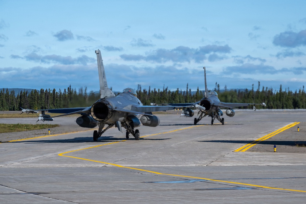 Two U.S. Air Force F-16 Fighting Falcon jet fighters are taxiing on a concrete runway apron, likely at a military airbase in Alaska based on the dense conifer tree line in the background. Both aircraft are armed with visible air-to-air missiles and external fuel tanks or munitions pods. The scene appears to be during daylight hours under partly cloudy skies.