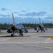 Two U.S. Air Force F-16 Fighting Falcon jet fighters are taxiing on a concrete runway apron, likely at a military airbase in Alaska based on the dense conifer tree line in the background. Both aircraft are armed with visible air-to-air missiles and external fuel tanks or munitions pods. The scene appears to be during daylight hours under partly cloudy skies.