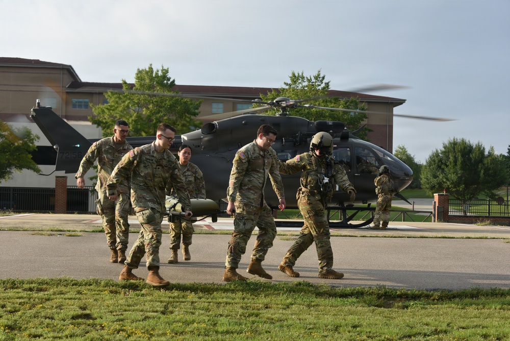 Military personnel in camouflage uniforms are carrying a patient on a litter/stretcher away from a landed military helicopter, likely during a medical evacuation exercise or operation. A flight crew member wearing a helmet is guiding the ground team. The scene takes place on a paved landing area near a multi-story building on what appears to be a military installation.