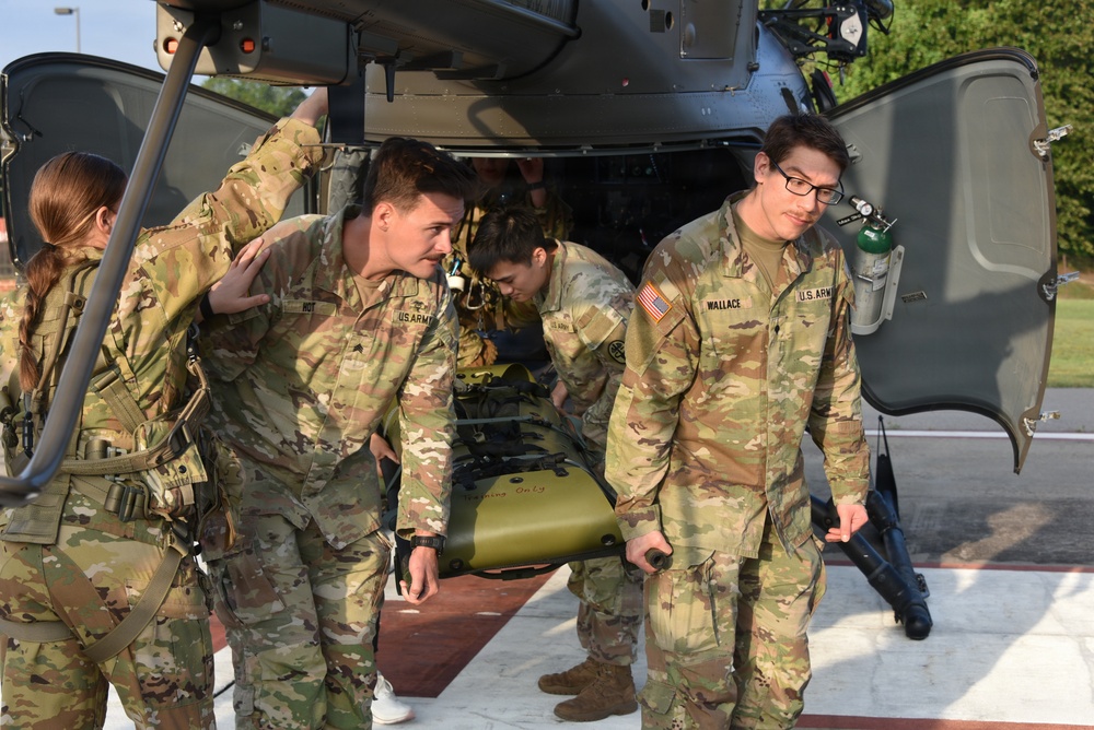 U.S. Army soldiers in OCP (Operational Camouflage Pattern) uniforms are conducting a medical evacuation (MEDEVAC) exercise, unloading a litter/stretcher from a military helicopter on what appears to be a helipad. The soldier on the right is identified by name tape as 'WALLACE' and is carrying a green oxygen cylinder. The scene depicts coordinated casualty evacuation training with multiple personnel working together around the aircraft.