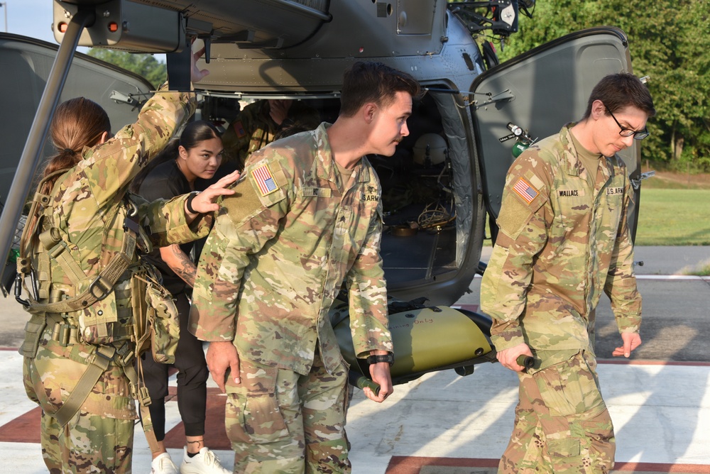 U.S. Army soldiers in OCP (Operational Camouflage Pattern) uniforms are conducting a medical evacuation (MEDEVAC) exercise near a military helicopter, likely a UH-72 Lakota or similar utility helicopter. Personnel are unloading or loading what appears to be a litter/stretcher from the aircraft on a marked helipad. One visible name tape reads 'WALLACE' and U.S. Army branch tapes and American flag patches are visible on multiple uniforms.