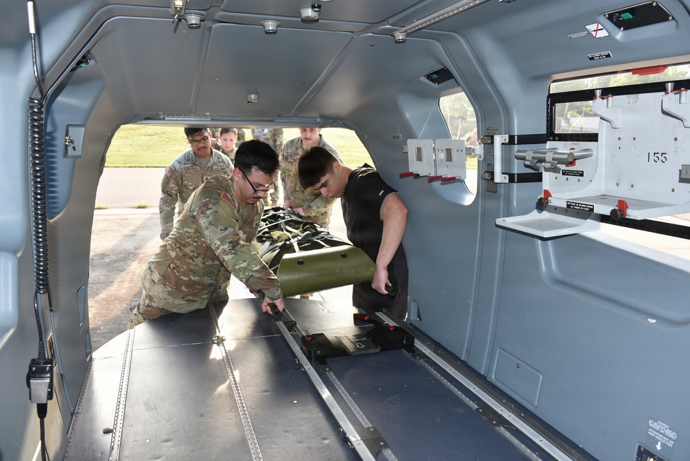 Military personnel in U.S. Army OCP (Operational Camouflage Pattern) uniforms are loading or positioning a green military litter/stretcher onto a rail system inside what appears to be a military medical or transport vehicle. Two soldiers are actively handling the litter while additional personnel observe from outside the vehicle. The interior of the vehicle is gray-painted metal with mounting hardware, rails, and instructional placards visible.