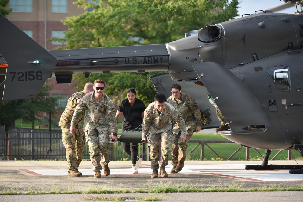 U.S. Army personnel are conducting a medical evacuation exercise or operation near a military helicopter on a helipad. Four soldiers in camouflage uniforms and one civilian in dark clothing are carrying a patient on a litter (stretcher) away from the aircraft. The helicopter, tail number 72156, is marked 'U.S. ARMY' and appears to be an HH-72 or similar utility/medevac helicopter.