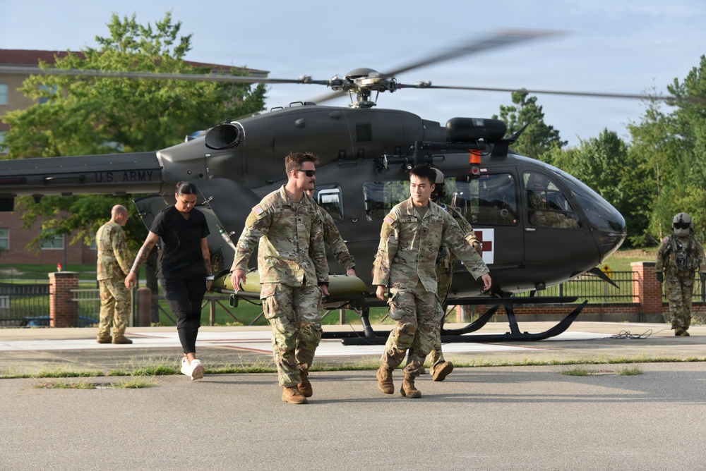 A U.S. Army medical helicopter (UH-72 Lakota) is parked on a landing pad with its rotors spinning. Several soldiers in camouflage uniforms are walking away from the aircraft, accompanied by a civilian in dark clothing. The scene appears to depict a medical evacuation or MEDEVAC training/operational scenario, evidenced by the red cross markings on the helicopter.