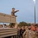 A U.S. Army soldier in OCP (Operational Camouflage Pattern) uniform stands elevated on the flatbed of a military truck, addressing a large formation of soldiers gathered below. The setting appears to be an early morning or dusk military installation, with personnel in both combat uniforms and Army PT (physical training) black shirts visible in the crowd. The scene suggests a pre-mission briefing, formation address, or training event.