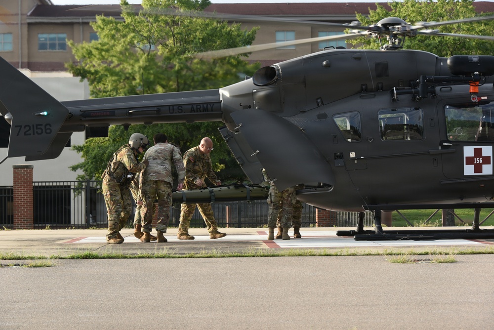 U.S. Army personnel in camouflage uniforms are loading or unloading a litter (stretcher) into a military medical evacuation helicopter marked with a red cross. The helicopter, tail number 72156, is identified as a UH-72 Lakota medevac aircraft bearing 'U.S. ARMY' markings. The scene appears to be a training exercise or actual medical evacuation operation on a designated helicopter landing pad near a military installation.