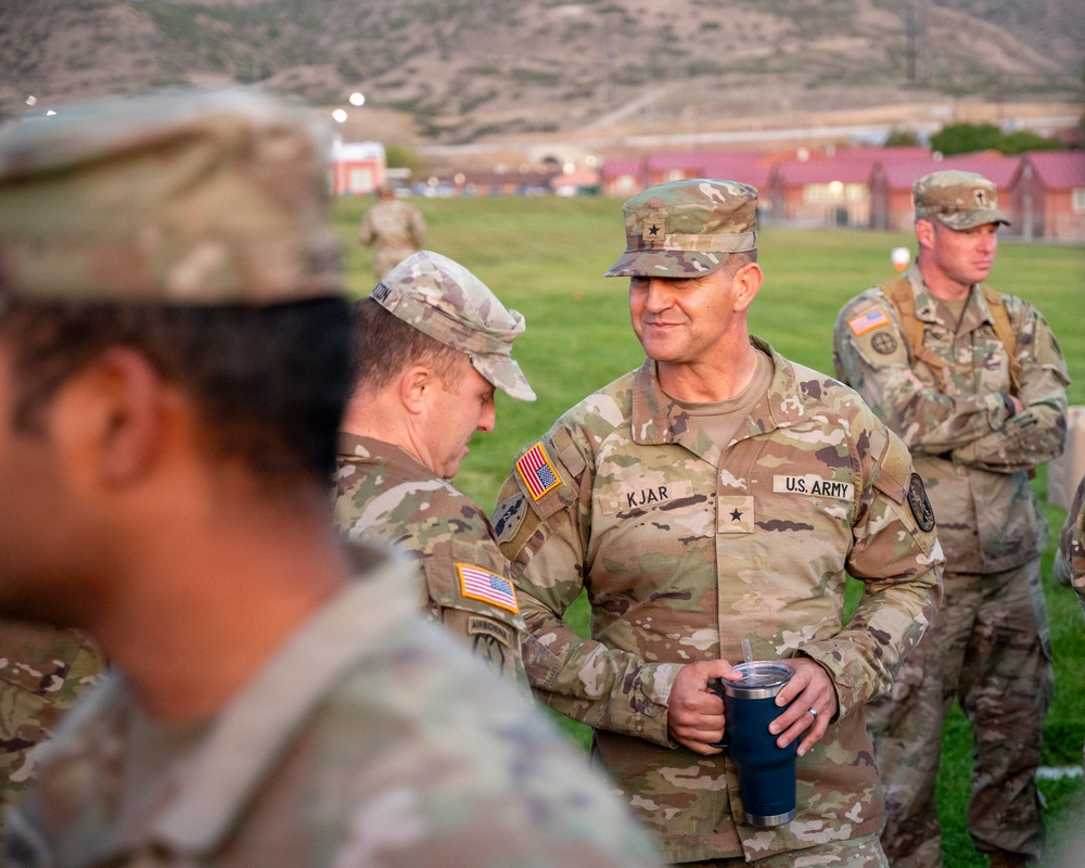 A group of U.S. Army soldiers are gathered outdoors on a grassy field, likely at a military installation. The central figure, identified by name tape as 'KJAR,' holds a dark tumbler cup and wears a single-star insignia indicating the rank of Brigadier General. The setting appears to be during early morning or evening hours based on the ambient lighting.