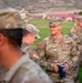 A group of U.S. Army soldiers are gathered outdoors on a grassy field, likely at a military installation. The central figure, identified by name tape as 'KJAR,' holds a dark tumbler cup and wears a single-star insignia indicating the rank of Brigadier General. The setting appears to be during early morning or evening hours based on the ambient lighting.