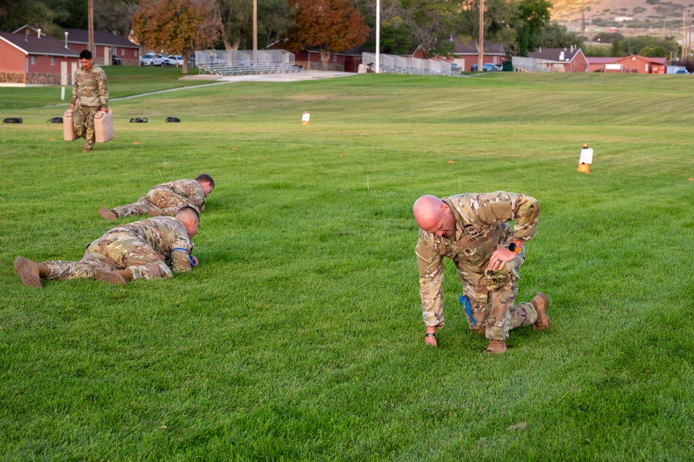 Military personnel in OCP (Operational Camouflage Pattern) uniforms are conducting physical training exercises on a grass field at what appears to be a military installation. Several soldiers are performing low crawl or push-up type movements on the ground, while one soldier in the background is carrying what appears to be a jerry can or weighted container. The setting includes red brick buildings, bleachers, and hills in the background, consistent with a U.S. Army base.
