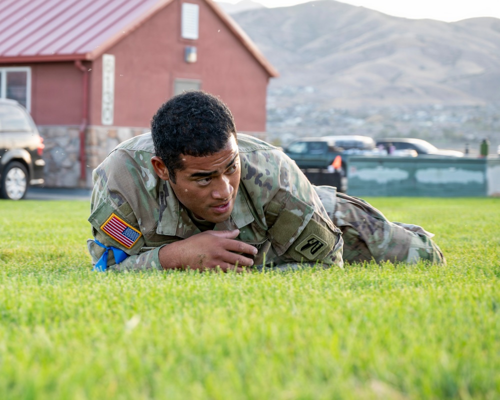 A U.S. Army soldier in OCP (Operational Camouflage Pattern) uniform is performing a low crawl exercise on a grass field. The soldier is wearing a U.S. flag patch on the left shoulder and appears to be engaged in a physical training or tactical movement drill. The setting appears to be a military installation with a red-roofed building and mountainous terrain visible in the background.