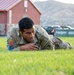 A U.S. Army soldier in OCP (Operational Camouflage Pattern) uniform is performing a low crawl exercise on a grass field. The soldier is wearing a U.S. flag patch on the left shoulder and appears to be engaged in a physical training or tactical movement drill. The setting appears to be a military installation with a red-roofed building and mountainous terrain visible in the background.