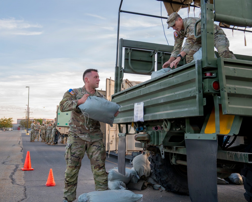 Military personnel in OCP (Operational Camouflage Pattern) uniforms are loading sandbags onto a military flatbed truck in what appears to be a staging area or parking lot. One soldier on the ground is handing a sandbag up to another soldier positioned on the truck bed. Additional uniformed personnel are visible in the background, suggesting a coordinated logistics or emergency preparedness operation.