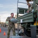 Military personnel in OCP (Operational Camouflage Pattern) uniforms are loading sandbags onto a military flatbed truck in what appears to be a staging area or parking lot. One soldier on the ground is handing a sandbag up to another soldier positioned on the truck bed. Additional uniformed personnel are visible in the background, suggesting a coordinated logistics or emergency preparedness operation.