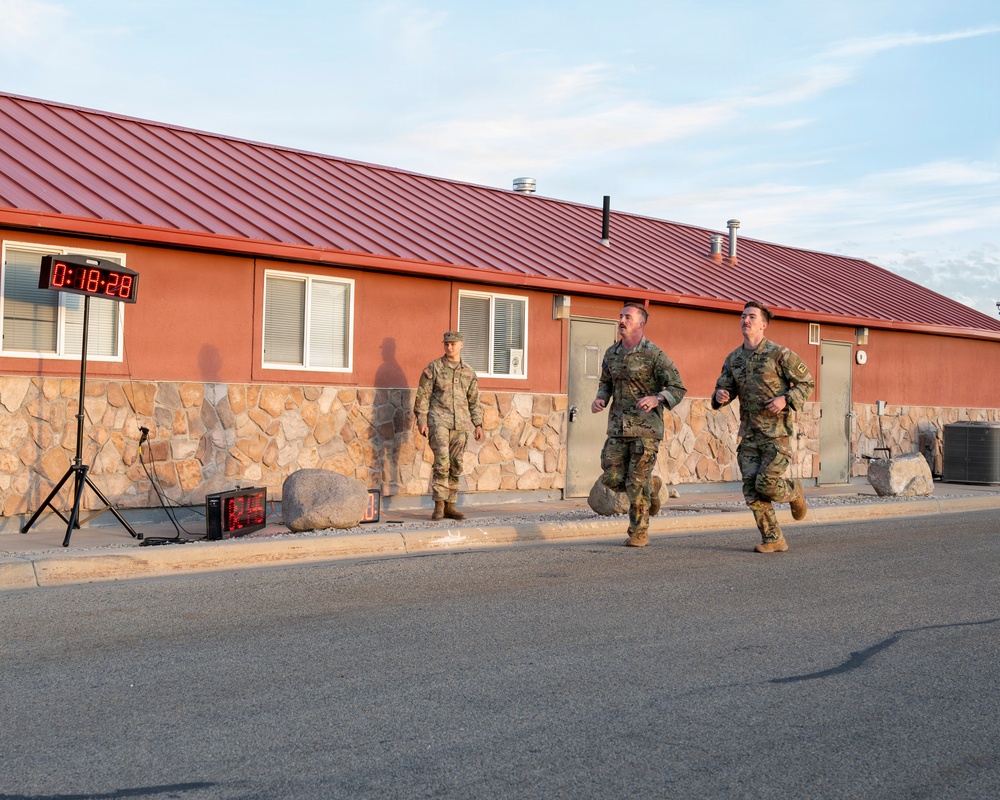 Two military personnel in camouflage uniforms are running past a building with a red metal roof and stone facade, while a third uniformed individual stands observing near the wall. A digital timer display on a tripod shows a time of 0:18:28, indicating this is a timed physical fitness test or run event. The setting appears to be a military installation during early morning or late afternoon based on the warm directional lighting.