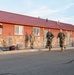 Two military personnel in camouflage uniforms are running past a building with a red metal roof and stone facade, while a third uniformed individual stands observing near the wall. A digital timer display on a tripod shows a time of 0:18:28, indicating this is a timed physical fitness test or run event. The setting appears to be a military installation during early morning or late afternoon based on the warm directional lighting.