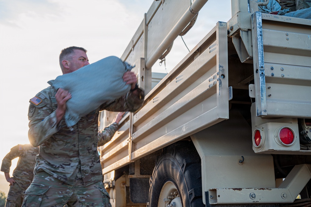 U.S. Army soldiers in MultiCam/OCP camouflage uniforms are loading or unloading sandbags from a military cargo truck, likely a LMTV or FMTV. The primary soldier in the foreground is carrying a large sandbag while another soldier assists in the background. This appears to be a flood control, disaster relief, or field fortification operation.