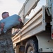 U.S. Army soldiers in MultiCam/OCP camouflage uniforms are loading or unloading sandbags from a military cargo truck, likely a LMTV or FMTV. The primary soldier in the foreground is carrying a large sandbag while another soldier assists in the background. This appears to be a flood control, disaster relief, or field fortification operation.
