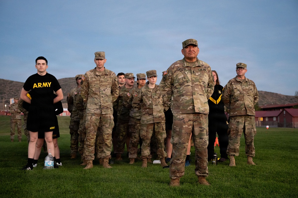 A group of U.S. Army personnel are standing at attention or in formation on a grass field, likely during a physical training or ceremonial event at a military installation. The group includes both uniformed soldiers in OCP (Operational Camouflage Pattern) uniforms and at least one individual in Army PT (physical training) attire. The setting appears to be early morning or evening based on the lighting and sky conditions.