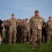 A group of U.S. Army personnel are standing at attention or in formation on a grass field, likely during a physical training or ceremonial event at a military installation. The group includes both uniformed soldiers in OCP (Operational Camouflage Pattern) uniforms and at least one individual in Army PT (physical training) attire. The setting appears to be early morning or evening based on the lighting and sky conditions.